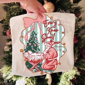 Puede incluir: Bolso tote crema con tem&aacute;tica navide&ntilde;a con un lazo rosa y el texto "Every Knee Shall Bow". El dise&ntilde;o incluye un &aacute;rbol de Navidad, el ni&ntilde;o Jes&uacute;s en un pesebre y Pap&aacute; Noel arrodillado. El bolso se sostiene frente a un &aacute;rbol de Navidad.