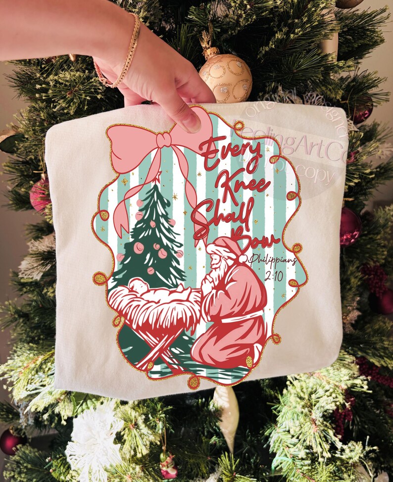 Puede incluir: Bolso tote crema con tem&aacute;tica navide&ntilde;a con un lazo rosa y el texto "Every Knee Shall Bow". El dise&ntilde;o incluye un &aacute;rbol de Navidad, el ni&ntilde;o Jes&uacute;s en un pesebre y Pap&aacute; Noel arrodillado. El bolso se sostiene frente a un &aacute;rbol de Navidad.