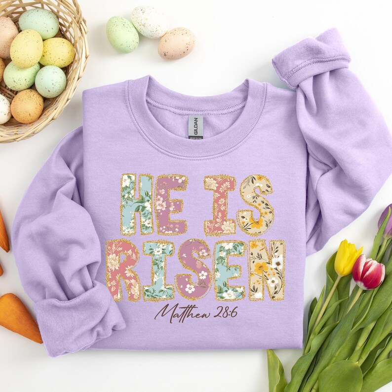 May include: A lavender sweatshirt with the words "He is Risen" in floral patterned letters. The text "Matthew 28:6" is printed below the floral letters.