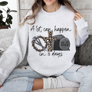 May include: A gray sweatshirt with a Christian Easter design. The design features a cross with a crown of thorns, nails, and a stone tomb with the text "He is Risen". The text "A lot can happen in 3 days" is also included.