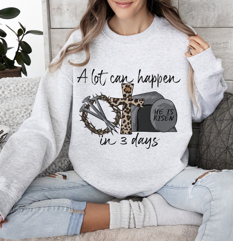 May include: A gray sweatshirt with a Christian Easter design. The design features a cross with a crown of thorns, nails, and a stone tomb with the text "He is Risen". The text "A lot can happen in 3 days" is also included.