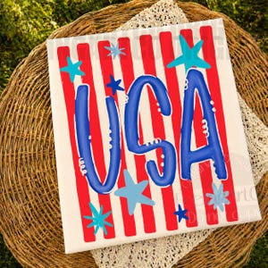 May include: A patriotic canvas art piece with a red and white striped background. The word "USA" is prominently displayed in blue, accented by blue stars. The artwork is placed on a woven basket with a lace trim.