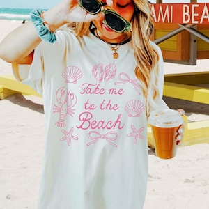 May include: Cream-colored t-shirt with the phrase "Take me to the Beach" in pink script, along with pink seashell, lobster, and starfish graphics. The shirt is worn with a pink cap and sunglasses. A beverage in a clear cup is held in the hand.