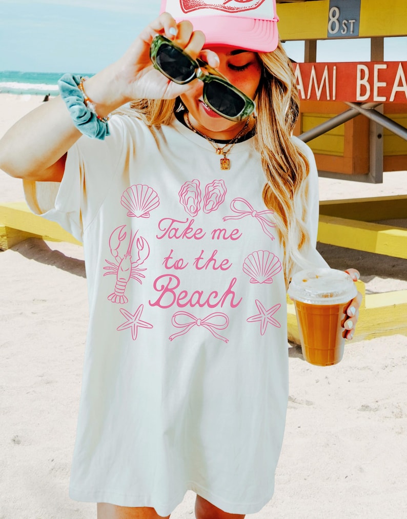 May include: Cream-colored t-shirt with the phrase "Take me to the Beach" in pink script, along with pink seashell, lobster, and starfish graphics. The shirt is worn with a pink cap and sunglasses. A beverage in a clear cup is held in the hand.