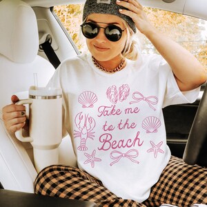 May include: A white t-shirt with the phrase "Take me to the Beach" in pink, along with seashell graphics. The person is wearing a gray beanie, sunglasses, and a gold chain. They are holding a white tumbler and wearing plaid pants.