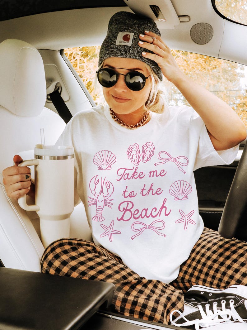 May include: A white t-shirt with the phrase "Take me to the Beach" in pink, along with seashell graphics. The person is wearing a gray beanie, sunglasses, and a gold chain. They are holding a white tumbler and wearing plaid pants.