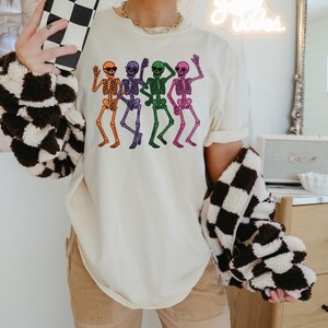Puede incluir: Camiseta color crema con un gr&aacute;fico de esqueletos bailando en naranja, morado, verde y rosa. Los esqueletos saludan y llevan gafas de sol. La camiseta se combina con pantalones color caqui y una chaqueta a cuadros blancos y negros.