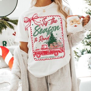 May include: White t-shirt with a festive graphic. The design features a red truck carrying books and a Christmas tree, with the text "Tis the Season to Read" in green and red. A red bow and decorative border complete the design.