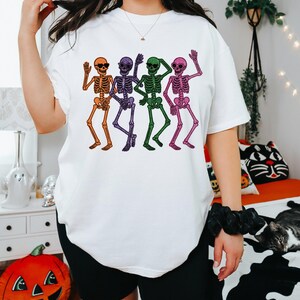 Puede incluir: Camiseta blanca con un gr&aacute;fico colorido de cuatro esqueletos bailando. Los esqueletos son de color naranja, morado, verde y rosa, con gafas de sol y brazos ondeando. La camiseta tiene un corte holgado y de gran tama&ntilde;o.
