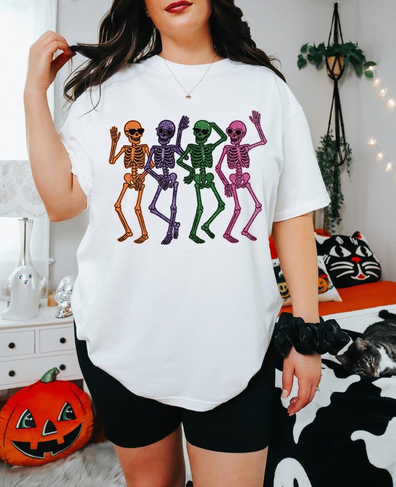Puede incluir: Camiseta blanca con un gr&aacute;fico colorido de cuatro esqueletos bailando. Los esqueletos son de color naranja, morado, verde y rosa, con gafas de sol y brazos ondeando. La camiseta tiene un corte holgado y de gran tama&ntilde;o.