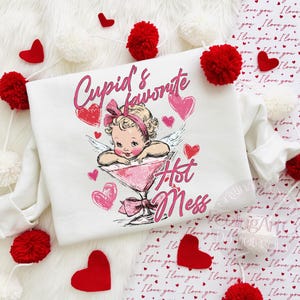 May include: White sweatshirt with a graphic of a vintage-style cupid in a martini glass, surrounded by pink hearts. The text reads "Cupid's Favorite Hot Mess". Red heart and pom-pom decorations are scattered around the sweatshirt.