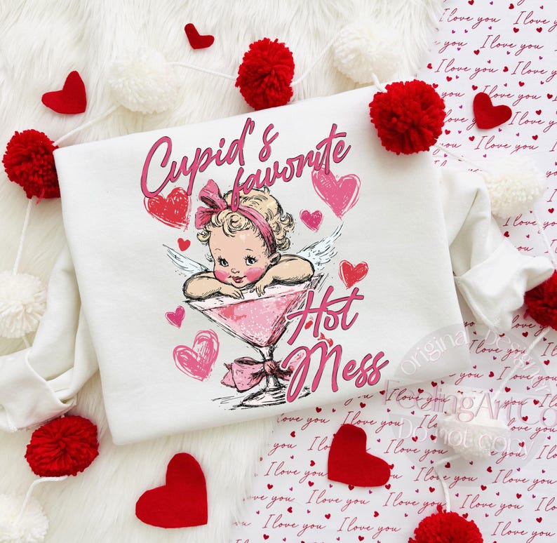May include: White sweatshirt with a graphic of a vintage-style cupid in a martini glass, surrounded by pink hearts. The text reads "Cupid's Favorite Hot Mess". Red heart and pom-pom decorations are scattered around the sweatshirt.