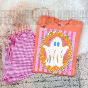 May include: An orange t-shirt featuring a white ghost graphic inside an orange and blue scalloped frame over pink vertical stripes, paired with pink and white gingham shorts and a gold-toned chain necklace with a circular pendant.
