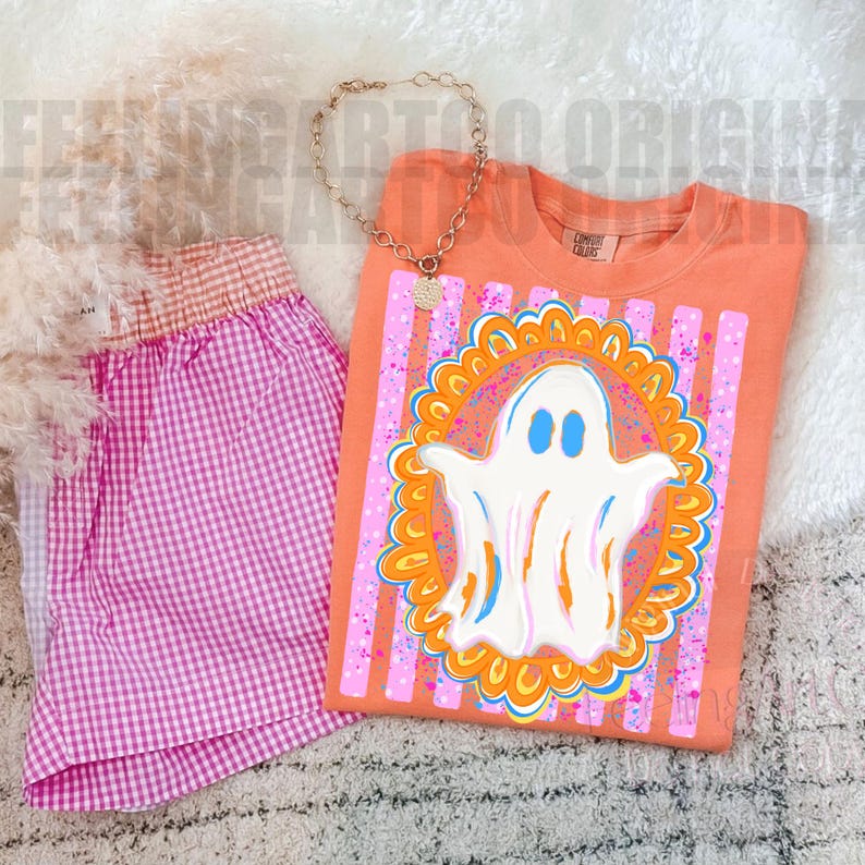 May include: An orange t-shirt featuring a white ghost graphic inside an orange and blue scalloped frame over pink vertical stripes, paired with pink and white gingham shorts and a gold-toned chain necklace with a circular pendant.
