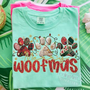 Puede incluir: Una camiseta verde claro con un dise&ntilde;o festivo "Merry Woofmas". El dise&ntilde;o presenta huellas de patas decoradas con patrones navide&ntilde;os y una guirnalda de luces. Una camiseta rosa est&aacute; apilada debajo.