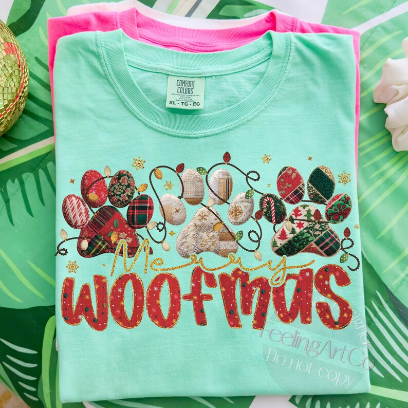 Puede incluir: Una camiseta verde claro con un dise&ntilde;o festivo "Merry Woofmas". El dise&ntilde;o presenta huellas de patas decoradas con patrones navide&ntilde;os y una guirnalda de luces. Una camiseta rosa est&aacute; apilada debajo.