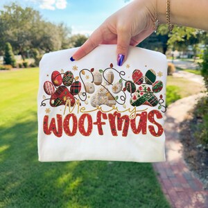 Puede incluir: Camiseta blanca con un dise&ntilde;o festivo que presenta huellas de patas de perro decoradas con motivos navide&ntilde;os y luces de cadena. El texto "Merry Woofmas" est&aacute; escrito en rojo y dorado. Una divertida camiseta tem&aacute;tica navide&ntilde;a para los amantes de los perros.