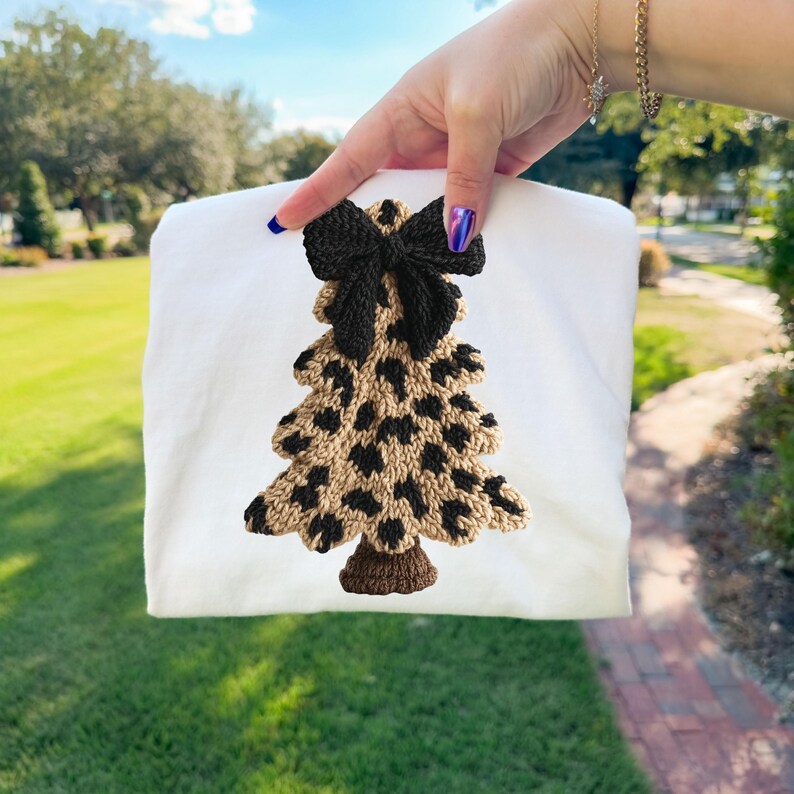 May include: A white sweatshirt featuring a knitted Christmas tree design. The tree has a leopard print pattern in tan and black, topped with a black bow. The tree trunk is brown.