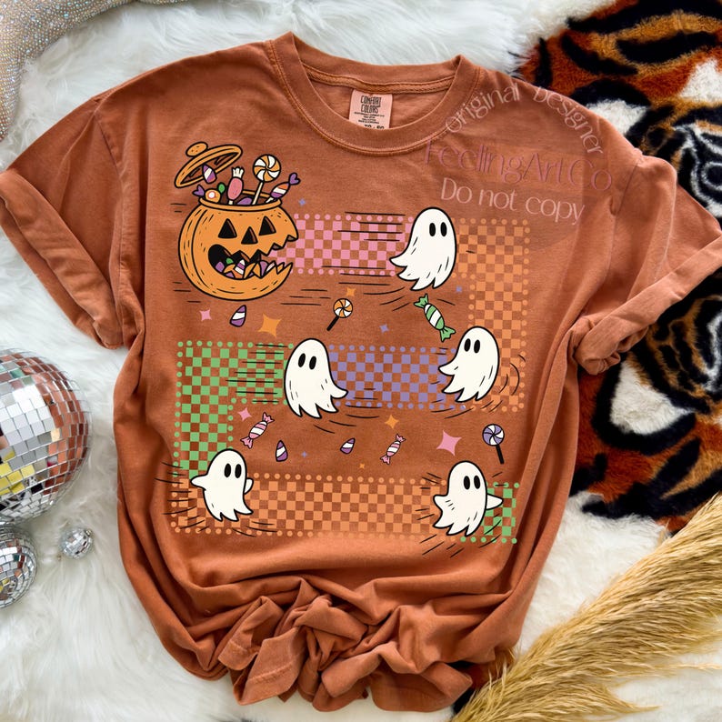May include: Burnt orange t-shirt featuring a Halloween-themed graphic. The design includes a jack-o'-lantern filled with candy, white ghosts, and colorful candies. The shirt has a classic crew neck and short sleeves. The text "FeelingArtCo Do not copy" is printed on the shirt.