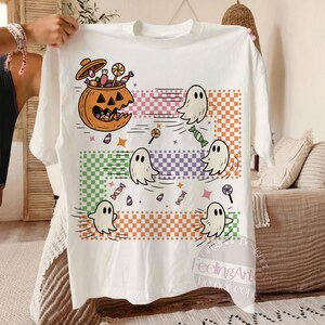 May include: White t-shirt featuring a Halloween-themed design. The graphic includes a jack-o'-lantern filled with candy, ghosts, and sweets. The design incorporates colorful checkered patterns and stars, creating a festive and playful look.