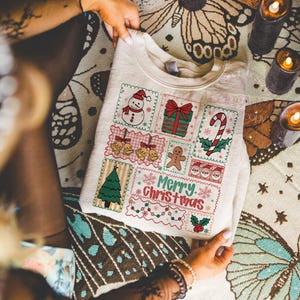 May include: Cream-colored sweatshirt featuring a festive Christmas design. The design includes a snowman, wrapped gift, candy cane, bells, gingerbread man, Christmas tree, and the words "Merry Christmas".