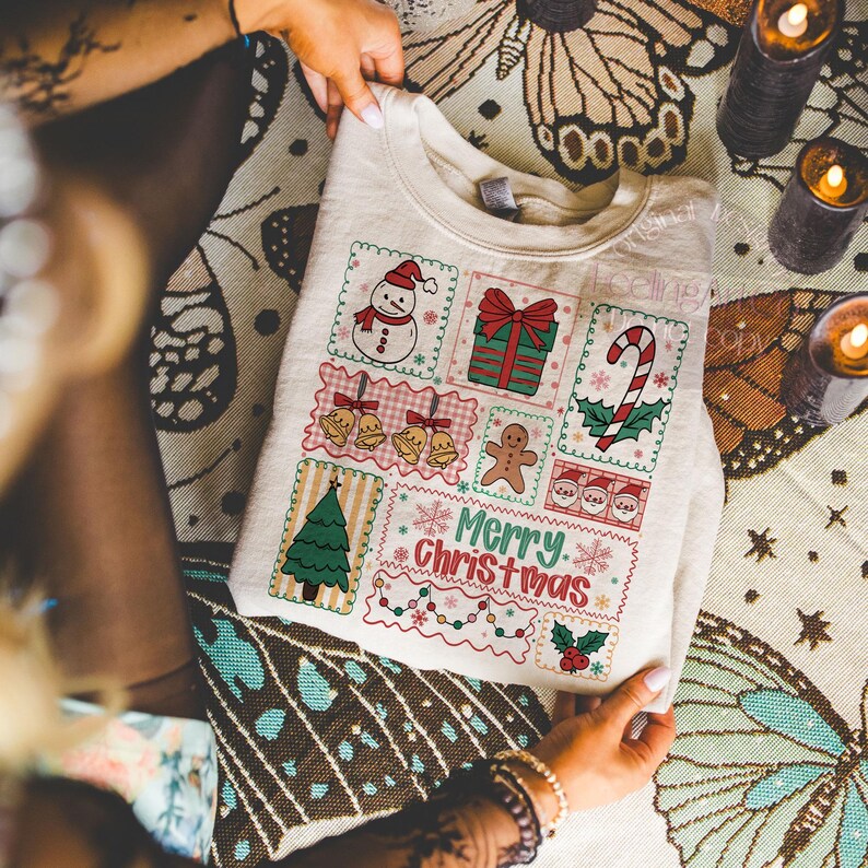 May include: Cream-colored sweatshirt featuring a festive Christmas design. The design includes a snowman, wrapped gift, candy cane, bells, gingerbread man, Christmas tree, and the words "Merry Christmas".
