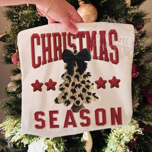 May include: A white Christmas-themed decorative pillow with the words "CHRISTMAS SEASON" in red embroidered lettering. The pillow features a leopard print Christmas tree and red star accents.