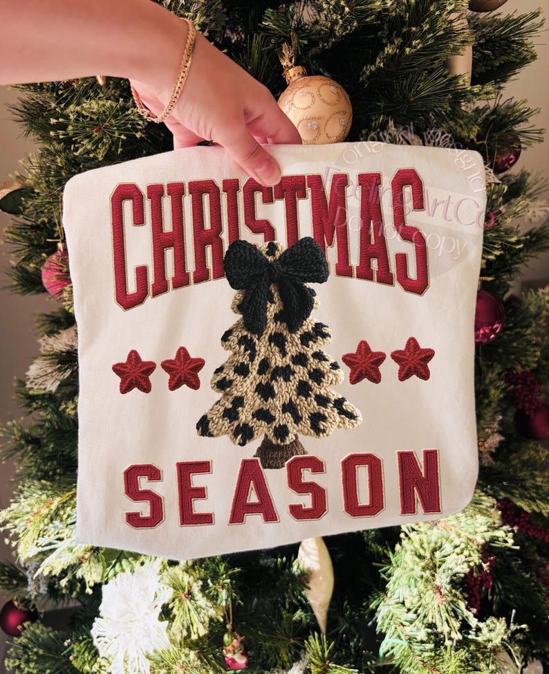 May include: A white Christmas-themed decorative pillow with the words "CHRISTMAS SEASON" in red embroidered lettering. The pillow features a leopard print Christmas tree and red star accents.