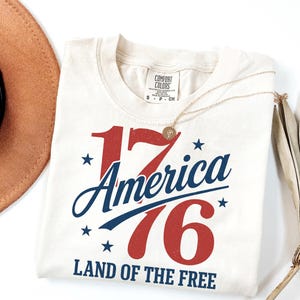 May include: Cream-coloured t-shirt with the text "1776 America" in red and blue, with stars and the phrase "Land of the Free." A gold necklace and a brown hat are also visible.