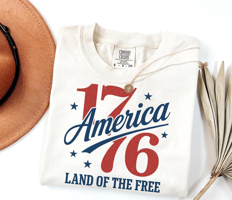 May include: Cream-coloured t-shirt with the text "1776 America" in red and blue, with stars and the phrase "Land of the Free." A gold necklace and a brown hat are also visible.