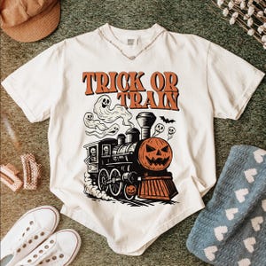 May include: Off-white t-shirt with a Halloween train graphic. The design includes a pumpkin face, ghosts, and the words "Trick or Train" in orange. The shirt is styled with accessories.