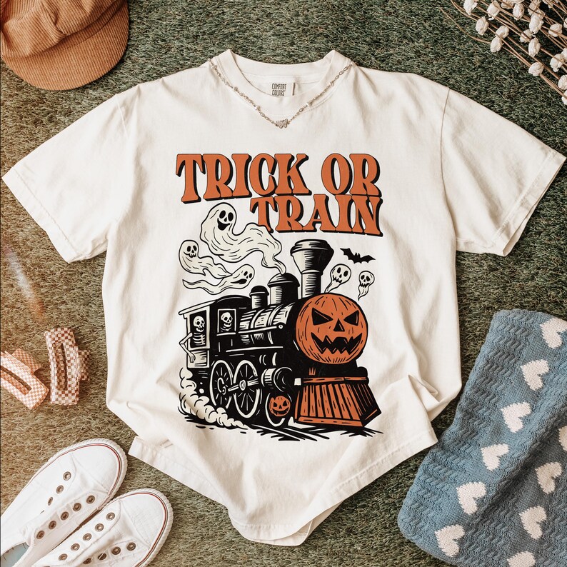 May include: Off-white t-shirt with a Halloween train graphic. The design includes a pumpkin face, ghosts, and the words "Trick or Train" in orange. The shirt is styled with accessories.