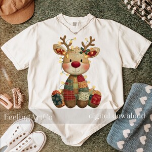 Puede incluir: Camiseta blanca con un dise&ntilde;o de reno con cuerpo de patchwork, cuernos y nariz roja. El reno est&aacute; adornado con luces de cadena. El texto "Feeling ArtCo" y "digital download" son visibles en la camiseta.