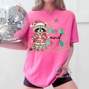 May include: A bright pink t-shirt featuring a raccoon wearing a Santa hat and a jacket adorned with Christmas lights. The text "Merry and Bright" is written in colorful letters. A disco ball is held in the background.
