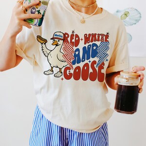 May include: A cream-coloured t-shirt with a cartoon goose wearing a baseball cap and holding a hot dog. The shirt features the text "Red, White, and Goose" with red, white, and blue accents. The person is wearing blue and white striped shorts.