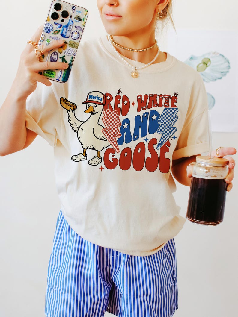 May include: A cream-coloured t-shirt with a cartoon goose wearing a baseball cap and holding a hot dog. The shirt features the text "Red, White, and Goose" with red, white, and blue accents. The person is wearing blue and white striped shorts.