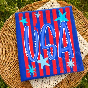 May include: A patriotic design featuring the word "USA" in blue, adorned with stars, set against a red and blue striped background. The design is on a blue rectangular surface.