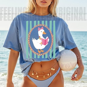 May include: Blue short-sleeved t-shirt featuring a cartoon goose reading a book, set within an oval frame with a scalloped edge. The goose wears a pink bow and holds a pencil. Vertical stripes in light blue and green are behind the oval.