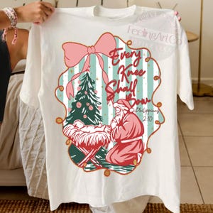 Puede incluir: Camiseta blanca con un dise&ntilde;o navide&ntilde;o. El dise&ntilde;o incluye un lazo rosa, un &aacute;rbol de Navidad y una representaci&oacute;n de Pap&aacute; Noel arrodillado. El texto "Every Knee Shall Bow" se muestra de forma destacada, junto con un vers&iacute;culo b&iacute;blico.