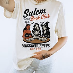 May include: Cream-colored t-shirt featuring the text "Salem Book Club" with illustrations of witches and bats. The shirt also includes the text "Massachusetts Est. 1692". The design is in black, red, and brown.