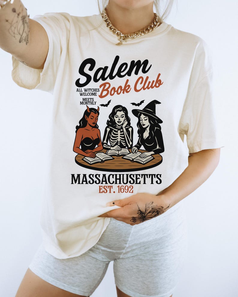 May include: Cream-colored t-shirt featuring the text "Salem Book Club" with illustrations of witches and bats. The shirt also includes the text "Massachusetts Est. 1692". The design is in black, red, and brown.