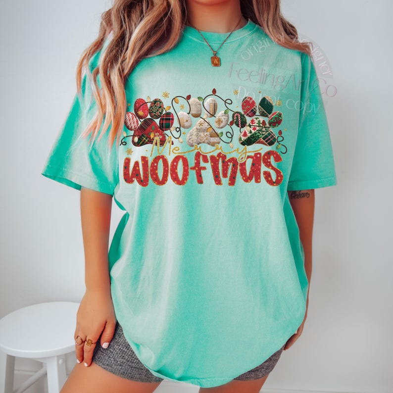Puede incluir: Camiseta turquesa con un dise&ntilde;o festivo. El dise&ntilde;o presenta huellas de patas, luces navide&ntilde;as y el texto "Merry Woofmas" en rojo. Las huellas de las patas est&aacute;n decoradas con cuadros y otros patrones.