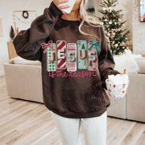 May include: A brown sweatshirt with the text "JESUS is the reason" in a colorful, festive design. The letters are decorated with plaid, stripes, and Christmas tree patterns, with bows on top. A cozy holiday-themed top.