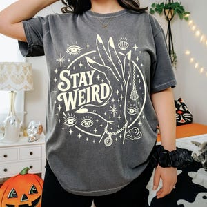 May include: Dark gray t-shirt with the words "Stay Weird" in a white, stylized font. The graphic includes a hand, eyes, stars, and a seashell. The shirt is short-sleeved and has a relaxed fit.