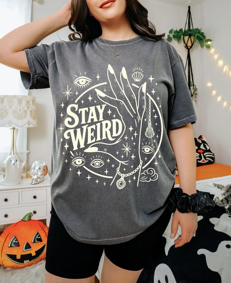 May include: Dark gray t-shirt with the words "Stay Weird" in a white, stylized font. The graphic includes a hand, eyes, stars, and a seashell. The shirt is short-sleeved and has a relaxed fit.