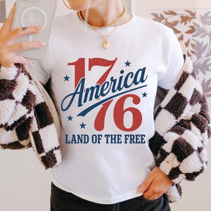 May include: White t-shirt featuring the text "17 America 76 Land of the Free" in red and blue, with star accents. The design has a vintage, patriotic feel. The person is wearing a black and white checkered jacket.