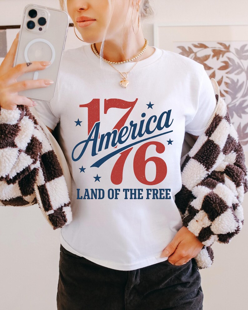 May include: White t-shirt featuring the text "17 America 76 Land of the Free" in red and blue, with star accents. The design has a vintage, patriotic feel. The person is wearing a black and white checkered jacket.