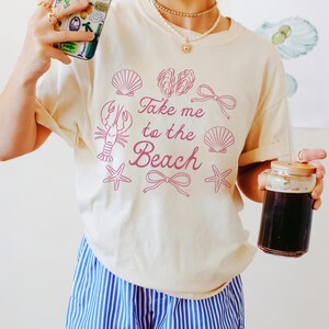 May include: Cream-colored t-shirt with the phrase "Take me to the Beach" in pink, along with seashell and lobster graphics. The person is wearing blue and white striped shorts and holding a phone with a beach-themed case and a glass of dark liquid.