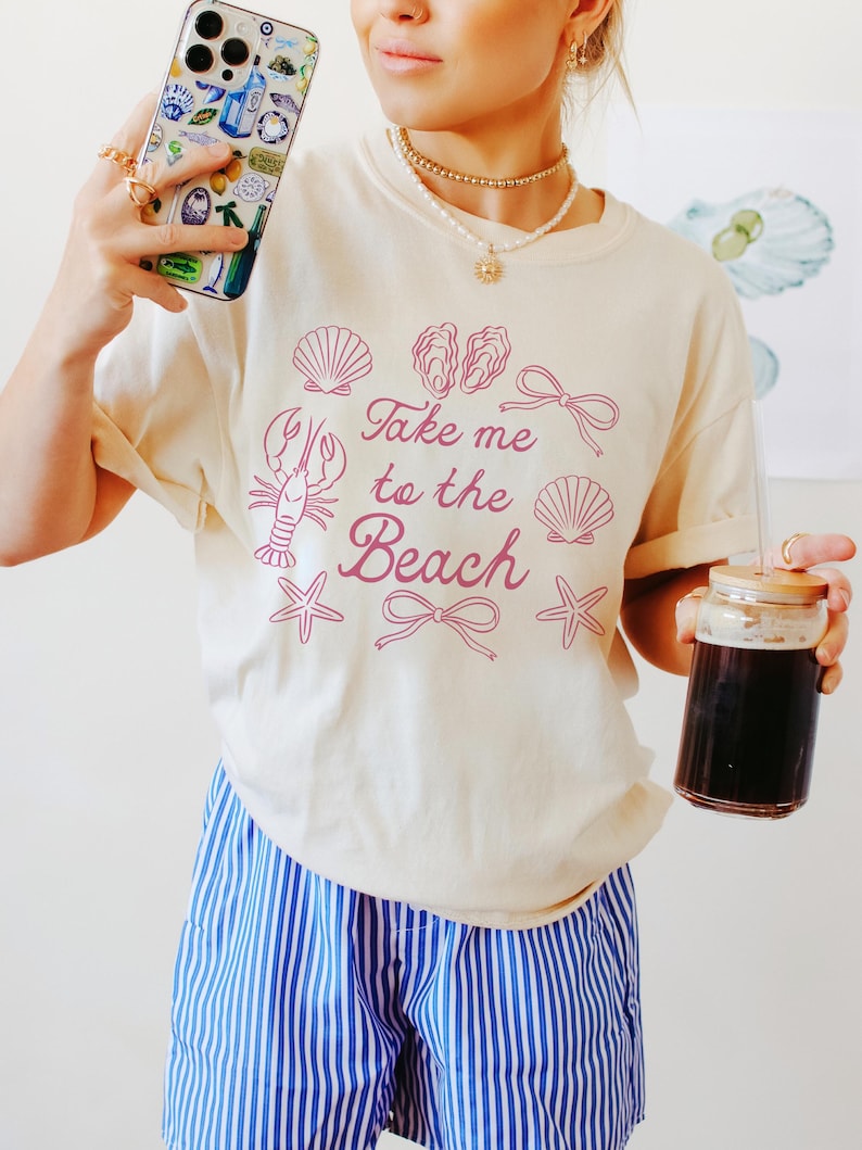 May include: Cream-colored t-shirt with the phrase "Take me to the Beach" in pink, along with seashell and lobster graphics. The person is wearing blue and white striped shorts and holding a phone with a beach-themed case and a glass of dark liquid.