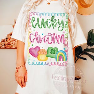 K&ouml;nnte beinhalten: Cremefarbenes T-Shirt mit den Worten "Lucky Charm" in Gr&uuml;n und Pink, umgeben von einem farbenfrohen Rahmen. Das Design umfasst ein Herz, einen Goldtopf, ein Kleeblatt und einen Regenbogen. Das Shirt hat eine l&auml;ssige, &uuml;bergro&szlig;e Passform.